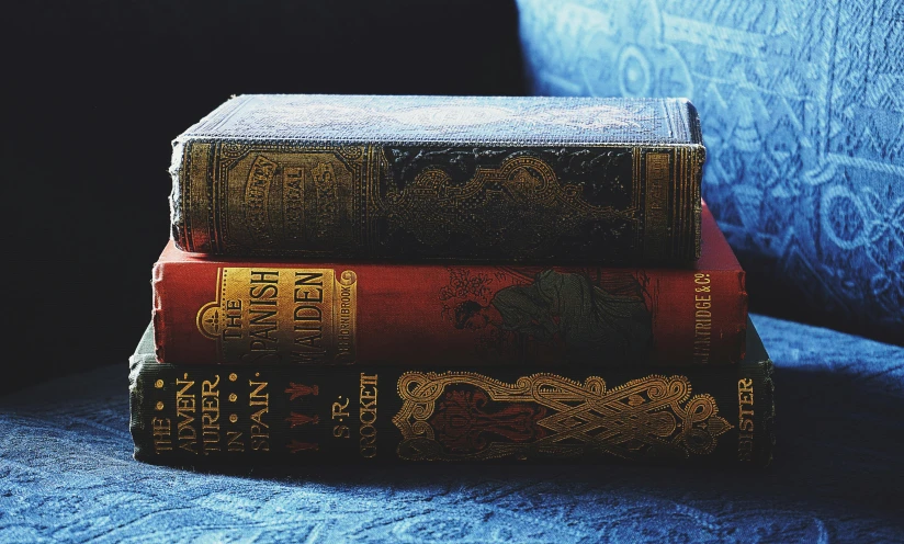 a stack of three books sitting on top of a blue couch, by Alice Mason, unsplash, romanticism, intricately detailed engravings, brown red blue, victorian textiles, red and blue back light