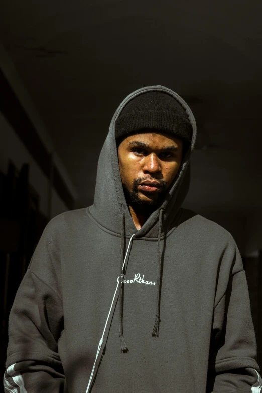 a man in a black hoodie standing in a dark room, by Bascove, frowning expression, ice cube, hat and hoodie, he is wearing a brown sweater