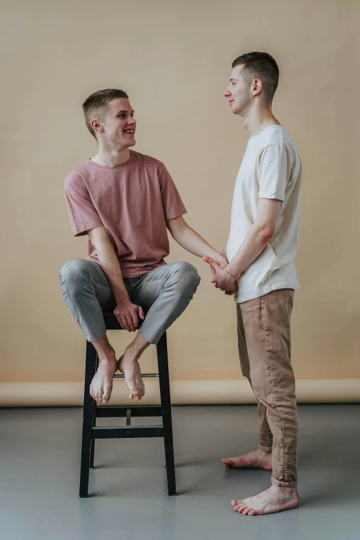 a man sitting on top of a chair next to another man, by Sara Saftleven, trending on pexels, teen boy, gentle calm doting pose, wearing pants, promotional image