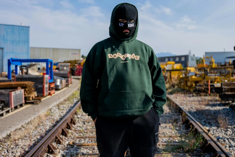 a person wearing a hoodie standing on a train track, by Bascove, wearing elaborate green and gold, official store photo, doran, debadged