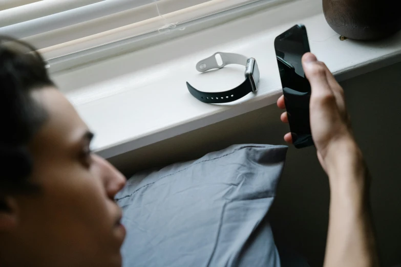 a close up of a person holding a cell phone, happening, sitting on the edge of a bed, futuristic product design, watched, next to a window