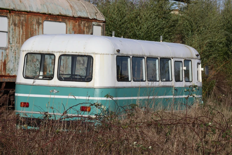 a blue and white bus parked next to a rusted building, overgrown with weeds, turquoise rust, dezeen, caravan