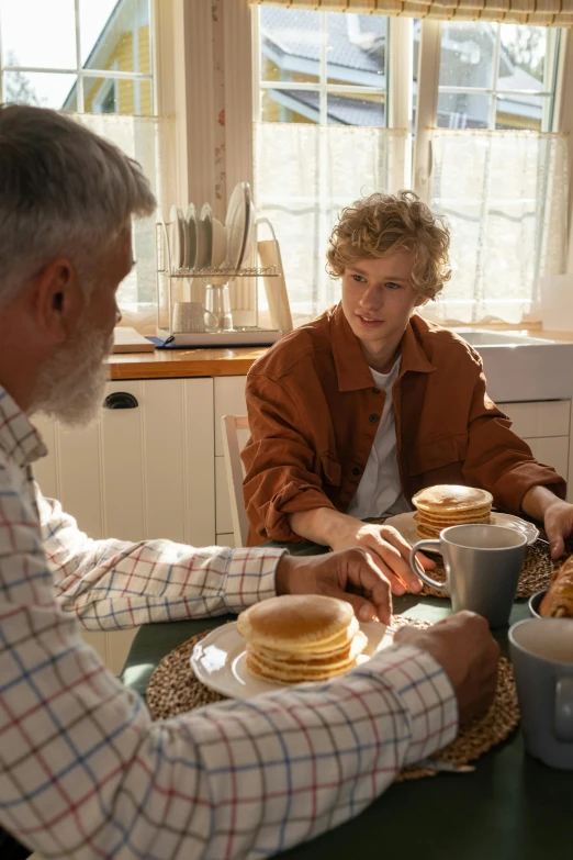 two men at a kitchen table eating pancakes and syrup