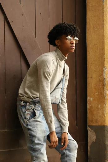 a man standing in front of a wooden door, an album cover, by Lily Delissa Joseph, trending on pexels, renaissance, portrait androgynous girl, wearing casual sweater, ( ( dark skin ) ), denim