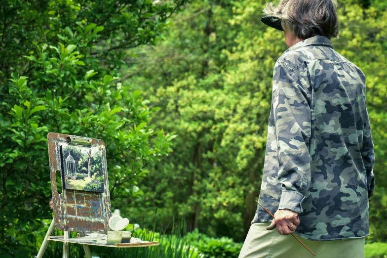 a man standing in front of a painting on a easel, inspired by Sir Alfred Munnings, visual art, enjoying a stroll in the forest, wearing camo, in the garden, close - up photograph
