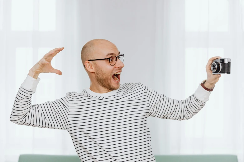 a man sitting on a couch taking a picture with a camera, an album cover, inspired by Leo Leuppi, pexels contest winner, antipodeans, arms spread wide, portrait of karl pilkington, jewish young man with glasses, waving hands