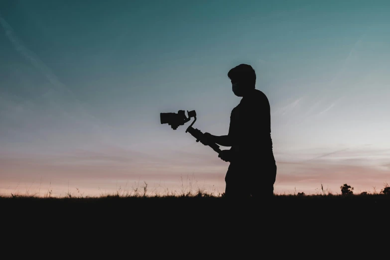 a man standing in a field holding a camera, unsplash contest winner, video art, silhouetted, screenwriter, a long-shot, over-the-shoulder shot