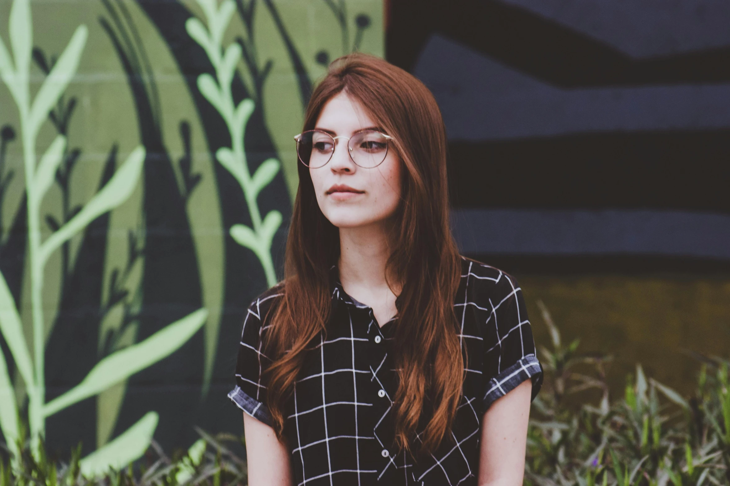 a woman sitting on a bench in front of a wall, a picture, inspired by Elsa Bleda, pexels contest winner, square rimmed glasses, chestnut hair, hipster, patterned