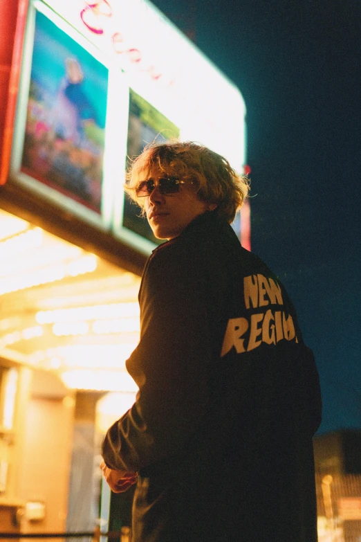 a man standing in front of a movie theater, an album cover, by Winona Nelson, trending on pexels, jamie campbell bower, wearing gold glasses, neon signs, rebirth