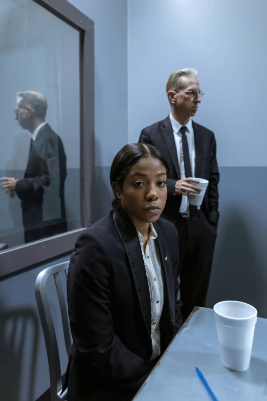 a woman sitting at a table in front of a mirror, two men in black, collateral, wearing causal black suits, prison scene