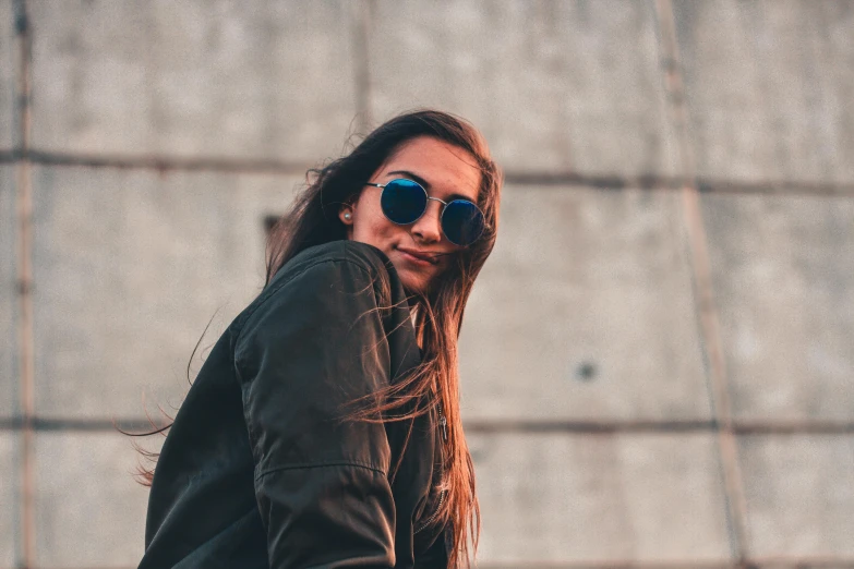a woman wearing sunglasses standing in front of a concrete wall, trending on pexels, happening, wearing an aviator jacket, happy girl, girl with long hair, unsplash 4k