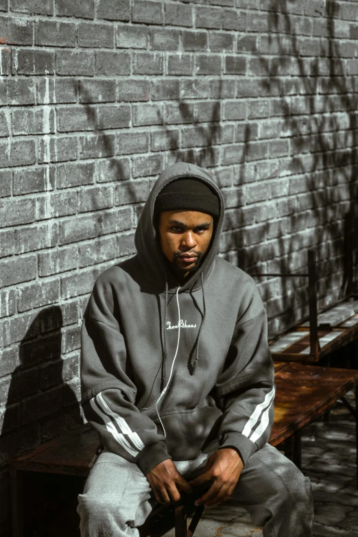 a man sitting on a bench in front of a brick wall, an album cover, trending on pexels, realism, wearing a grey hooded sweatshirt, african canadian, sythwave, serious look