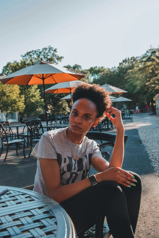 a woman sitting at a table with an umbrella in the background, a portrait, by Lily Delissa Joseph, trending on unsplash, tan skin a tee shirt and shorts, natural hair, androgynous male, park in background