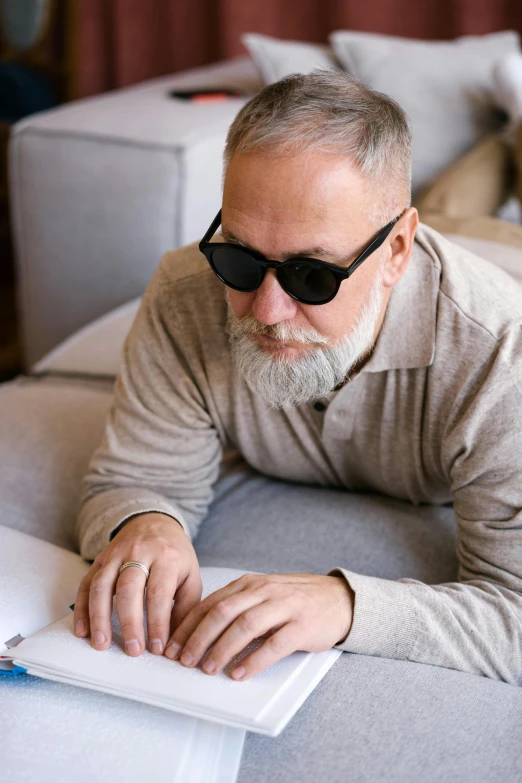 a man sitting on a couch reading a book, inspired by Carl Gustaf Pilo, implanted sunglasses, gray hair and beard, profile image, compassionate