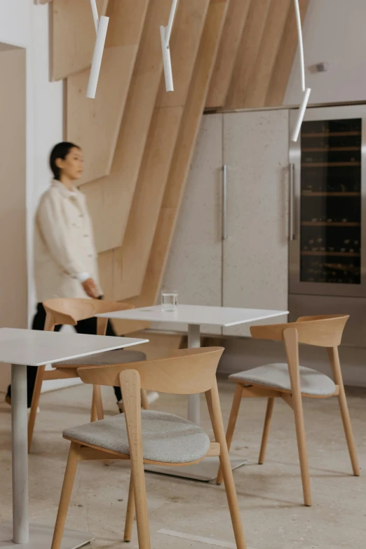 a woman standing in a kitchen next to a table, inspired by Barthélemy Menn, trending on unsplash, conceptual art, john pawson, cafe interior, koji morimoto shinjuku, chairs