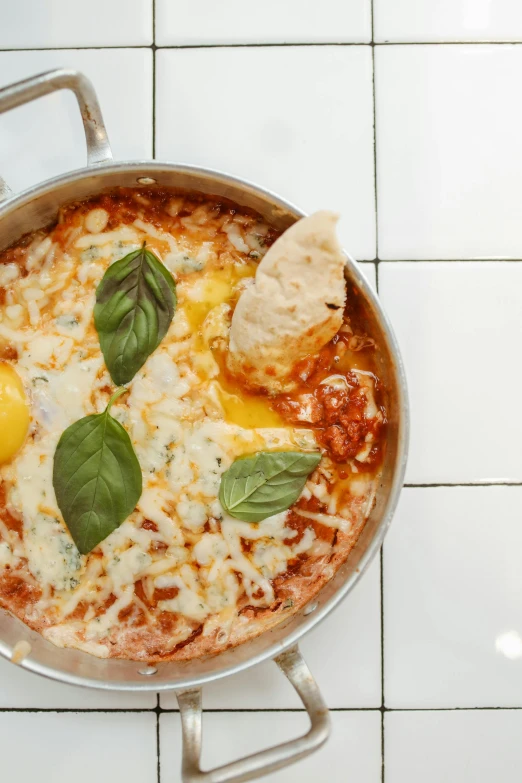 a pan filled with food sitting on top of a white tiled floor, by Anita Malfatti, eggs, pizza on a table, basil, lasagna