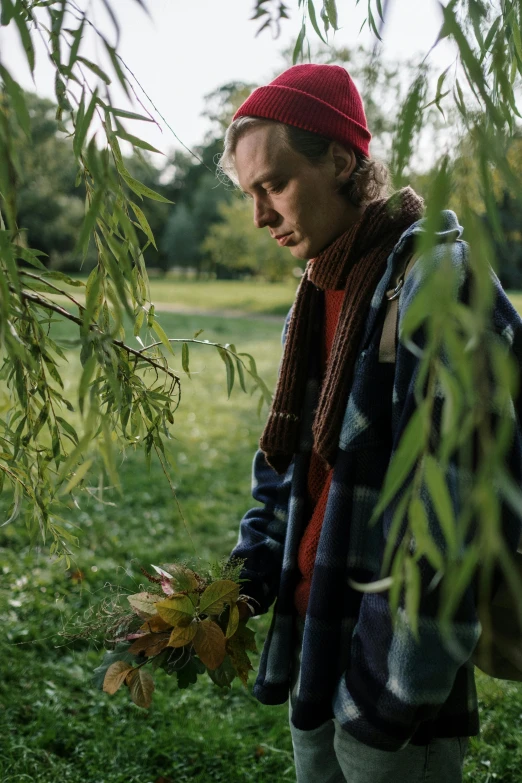 a man standing under a tree holding a bunch of leaves, by Penelope Beaton, unsplash, visual art, movie still of a tired, jamie campbell bower, he‘s wearing a red scarf, gardening