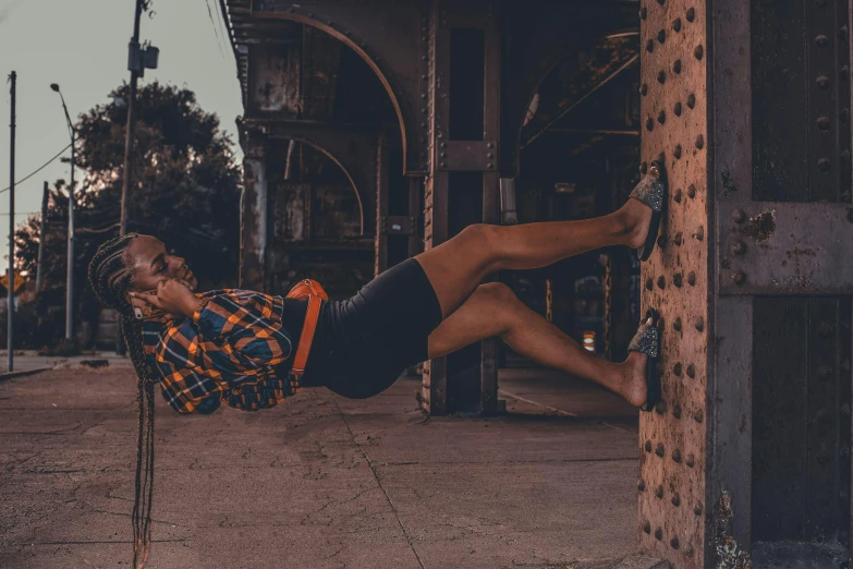 a woman laying on a pole in front of a building, by Ingrida Kadaka, pexels contest winner, happening, ( ( dark skin ) ), bra and shorts streetwear, sitting under bridge, queer woman