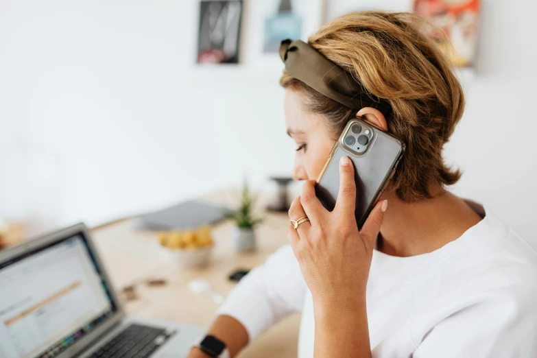 a woman talking on a cell phone next to a laptop, by Julia Pishtar, trending on pexels, hair tied in a ponytail, cell cover style, phone!! held up to visor, worksafe. instagram photo