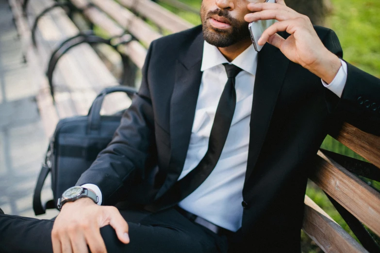 a man sitting on a bench talking on a cell phone, trending on pexels, black luxurious suit, long tie, raphael lecoste, security agent