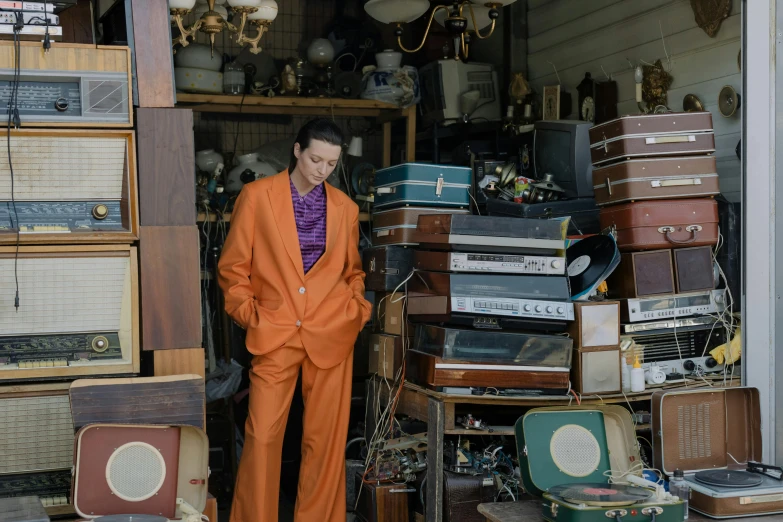a man in an orange suit standing in front of a pile of suitcases, an album cover, by Winona Nelson, unsplash, maximalism, standing in his cluttered garage, julian ope, jean deville, bella poarch
