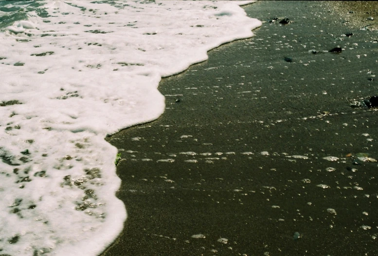 a person riding a surfboard on top of a sandy beach, an album cover, by Elsa Bleda, unsplash, wave of water particles, black sand, 1990's photo, beach is between the two valleys