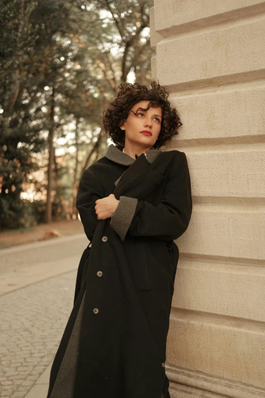 a woman leaning against a wall in a black coat, by Lucia Peka, pexels contest winner, renaissance, curly and short top hair, roma, wearing hunter coat, rectangle