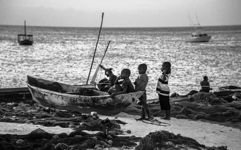 a group of people standing on top of a beach next to a boat, a black and white photo, by John Hutton, pexels, kids playing, masai, fishing boats, evening time