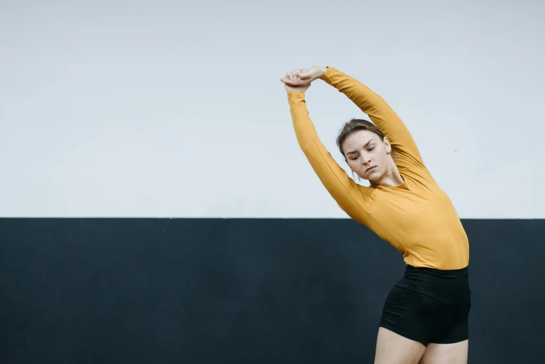 a woman in a yellow shirt and black shorts stretching her arms, unsplash, arabesque, background image, performance, wearing leotard, college