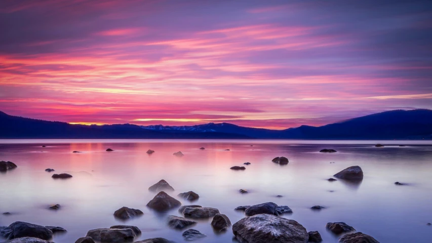 a sunset over a body of water with rocks in the foreground, inspired by Edwin Georgi, unsplash contest winner, pink and purple, lakeside mountains, multiple stories, calm vivid colors