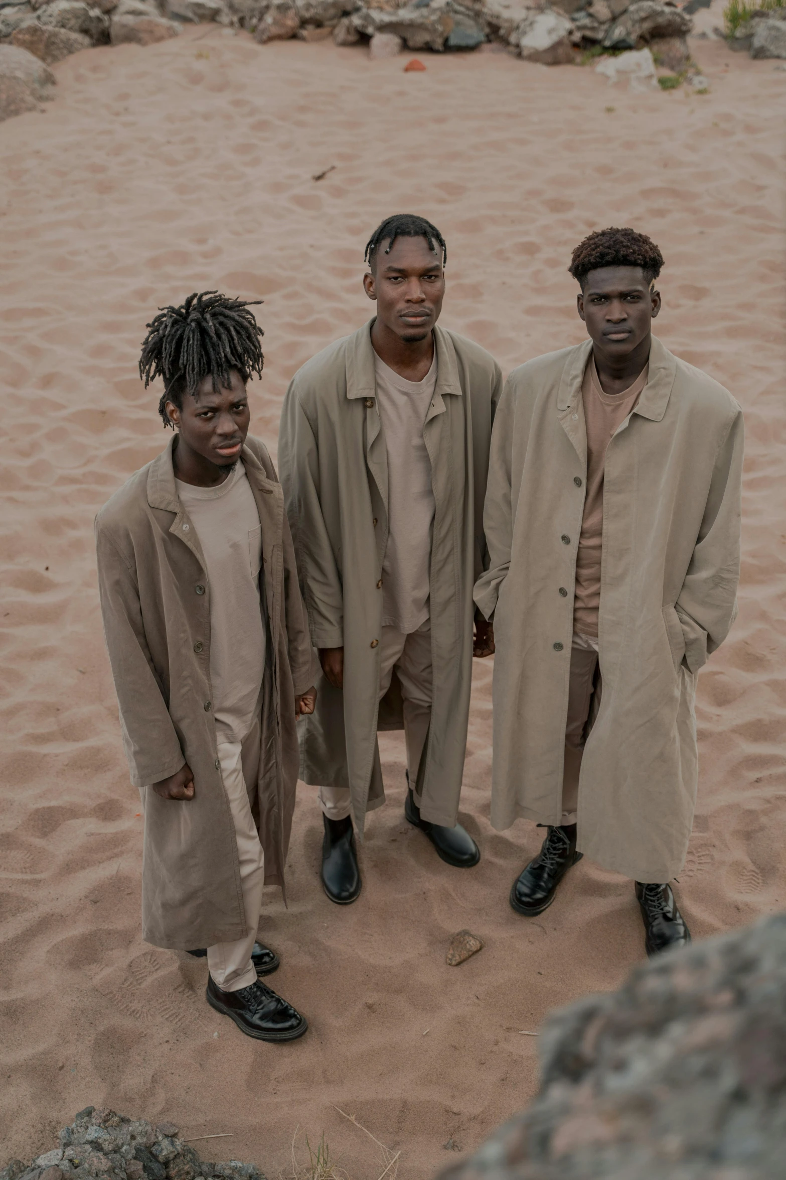 a group of men standing on top of a sandy beach, an album cover, by Caro Niederer, trending on pexels, afrofuturism, trench coat, taupe, ( ( theatrical ) ), himba hairstyle