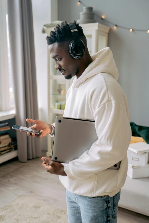 a man standing in a living room holding a laptop and headphones, trending on pexels, in white turtleneck shirt, black man, thumbnail, earbuds