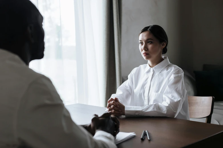 a man and a woman sitting at a table, wearing a white shirt, mental health, female lawyer, reaching out to each other