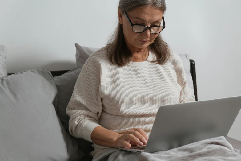 a woman sitting on a bed using a laptop computer, pexels, going gray, wearing small round glasses, avatar image, grey