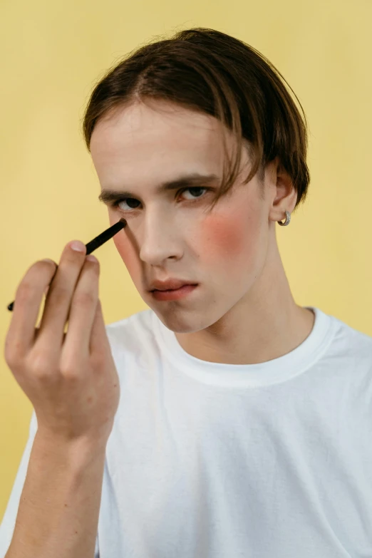 a man in a white shirt is doing makeup, inspired by Mac Conner, trending on pexels, teen boy, complimentary eyeliner, nonbinary model, holding pencil