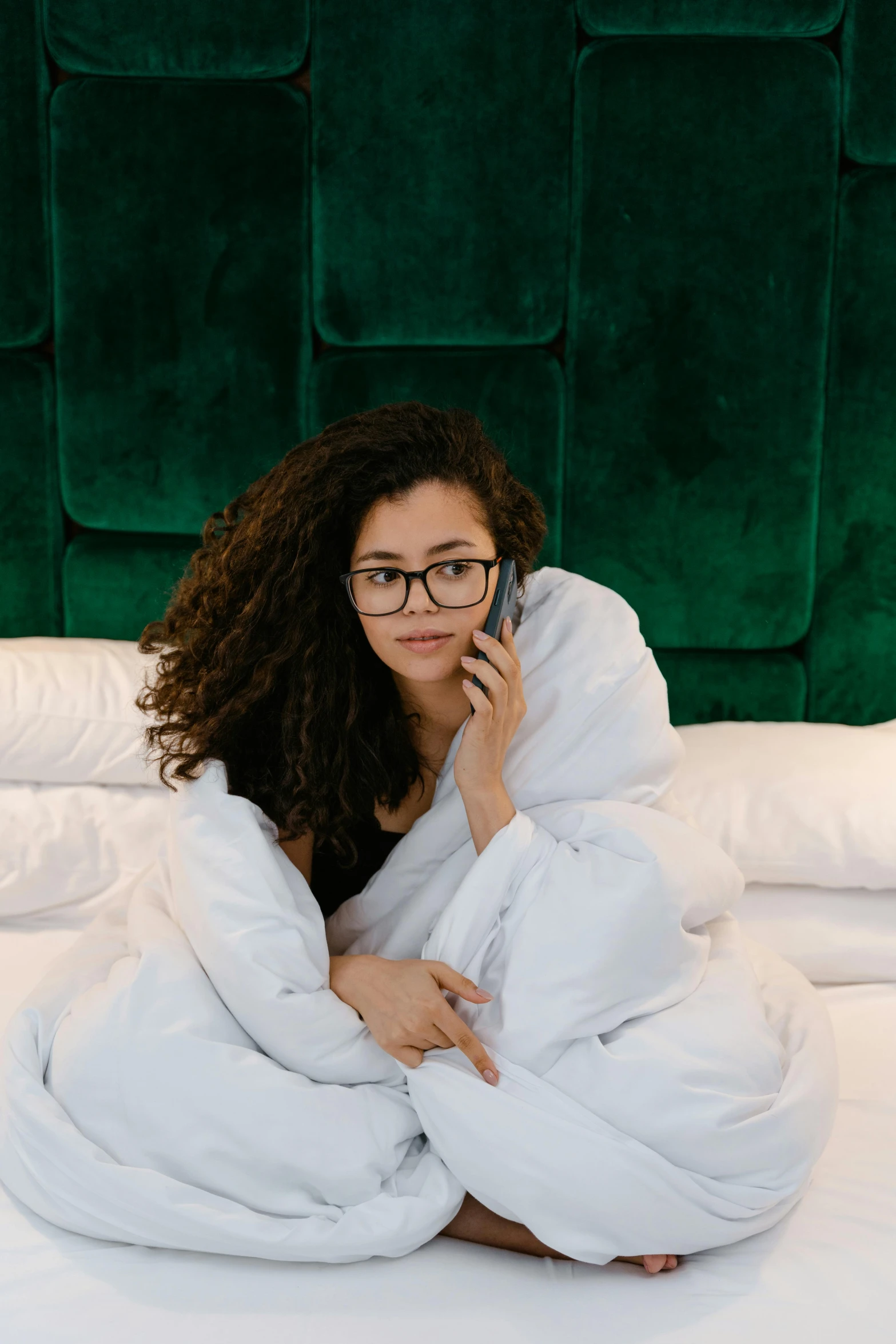 a woman sitting on a bed talking on a cell phone, curly haired, wavy long black hair and glasses, curled up under the covers, 2019 trending photo