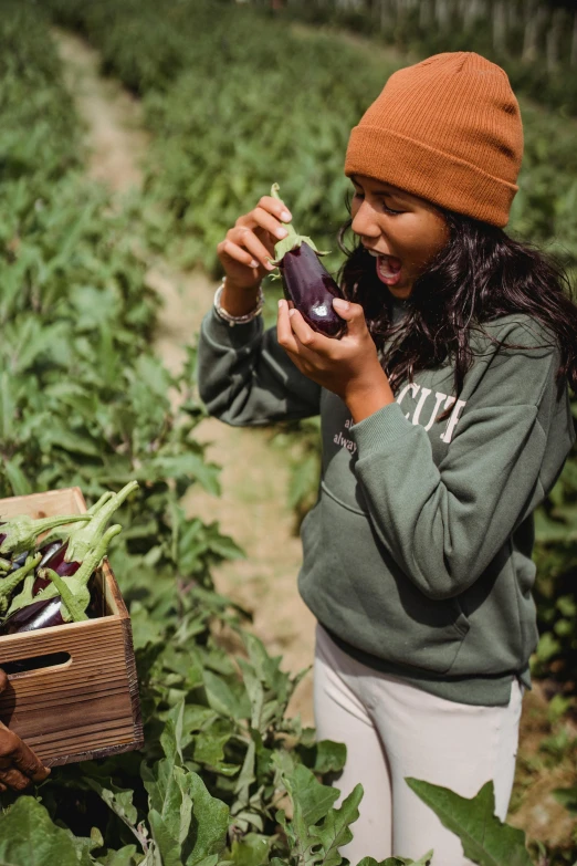 a woman standing in a field holding an eggplant, pexels contest winner, inspect in inventory image, black haired girl wearing hoodie, wearing farm clothes, performing