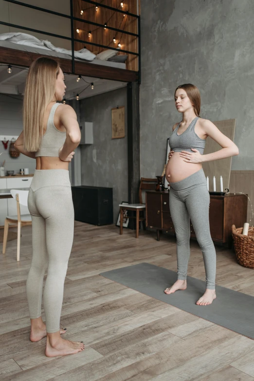 a pregnant woman standing on yoga mat next to a woman in the room