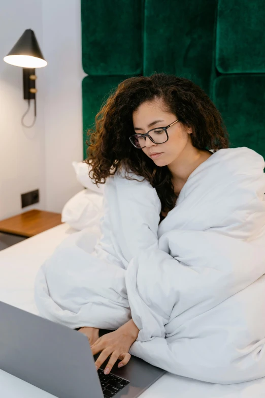 a woman sitting on a bed using a laptop, trending on pexels, happening, wearing nanotech honeycomb robe, wearing black rimmed glasses, sleepy expression, curly haired