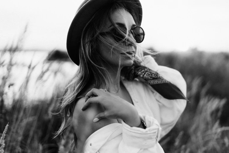 a black and white photo of a woman in a field, trending on pexels, wearing sunglasses and a hat, long windy hair style, young sexy elegant woman, wearing a white blouse