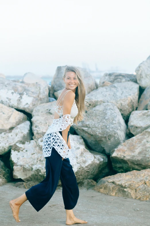 a woman walking on a beach next to large rocks, a picture, unsplash, happening, full body! pretty face, with long blond hair, smiling and dancing, white lace clothing