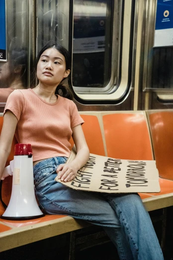 a woman sitting on a train holding a sign, trending on reddit, feminist art, 8 0 s asian neon movie still, ny, tan, 2019 trending photo
