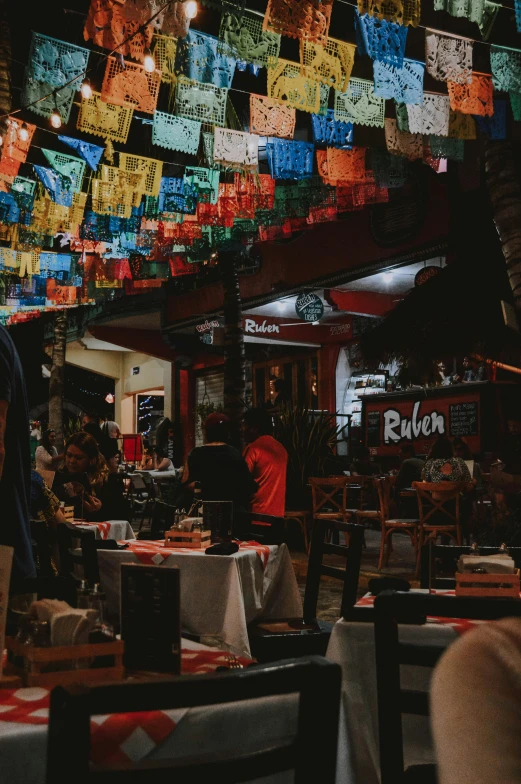 a group of people sitting at tables in a restaurant, spanish alleyway, red and black flags waving, colorful projections, chilaquiles