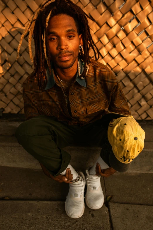 a man with dreadlocks sitting on the ground, by Kurt Trampedach, holding a gold bag, skateboarder style, hand holding cap brim, headshot