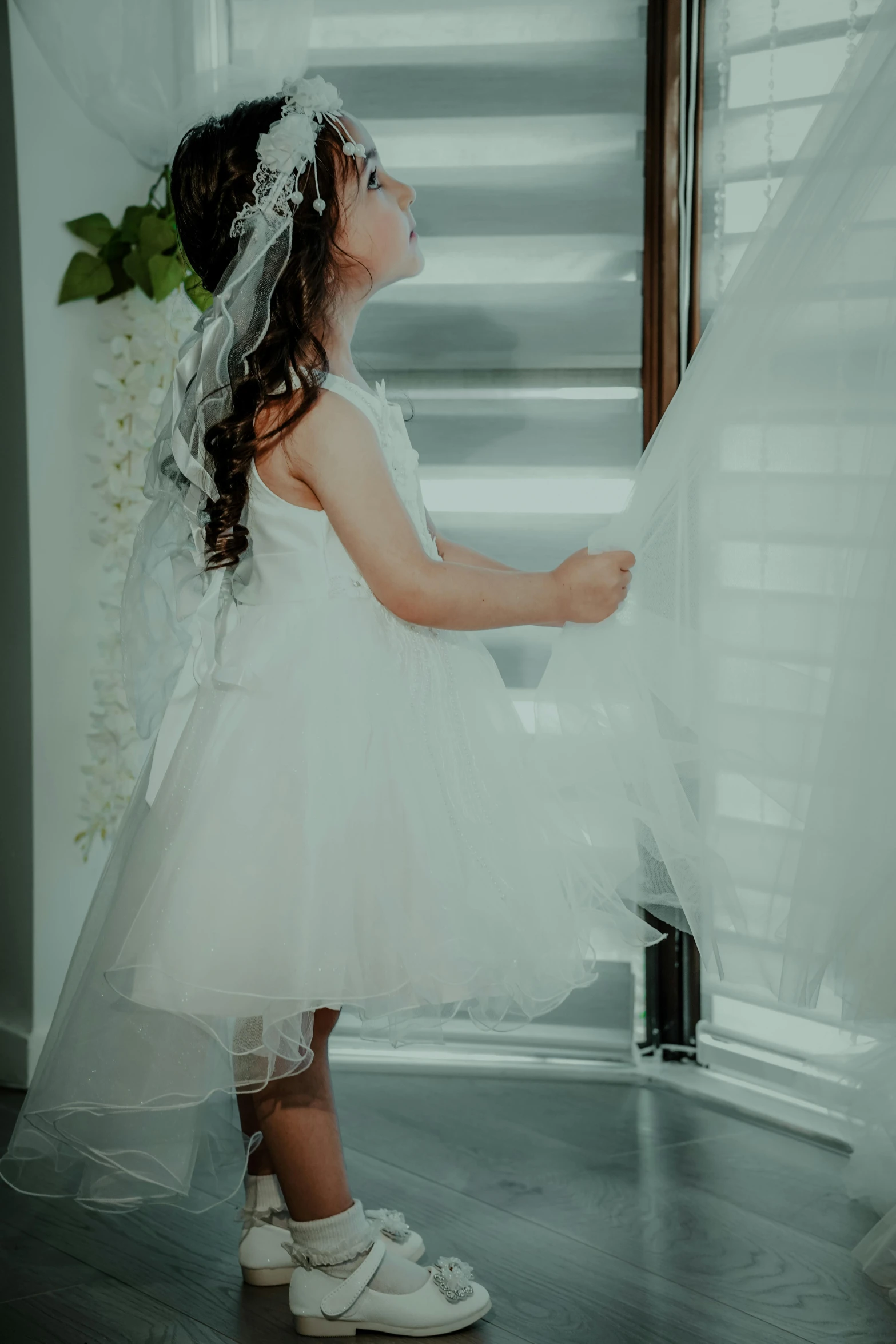 a little girl standing in front of a window, wearing a wedding dress, shot from the side, angel themed