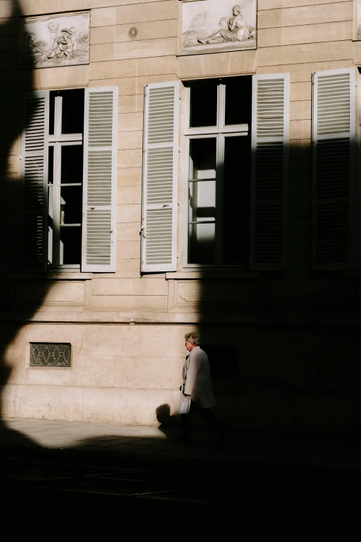 a man sitting on the sidewalk in front of a building, pexels contest winner, paris school, half in shadow, shutters, walking away from camera, medium format. soft light