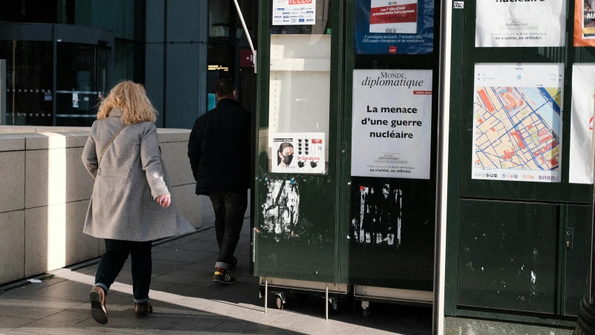 a woman walking down a sidewalk next to a tall building, a poster, les automatistes, emmanuel macron, post-nuclear, ap news photograph, a bald