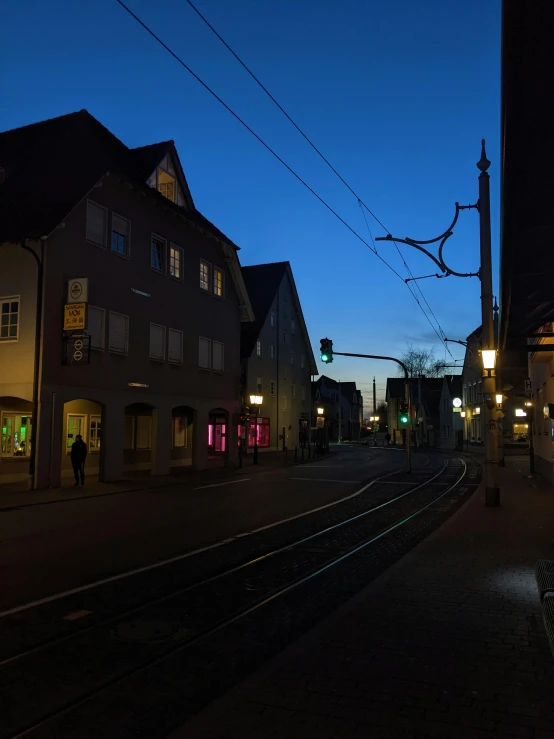 a train traveling down a train track next to a tall building, a picture, by Kristian Zahrtmann, street lanterns glow, detmold, nightfall. quiet, house's and shops and buildings