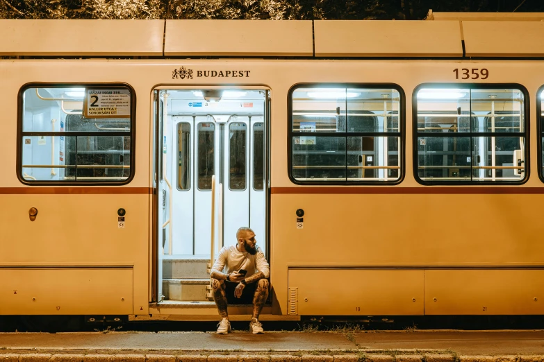 a man sitting on a bench in front of a train, by Matija Jama, pexels contest winner, happening, street tram, yellow street lights, brown, thumbnail