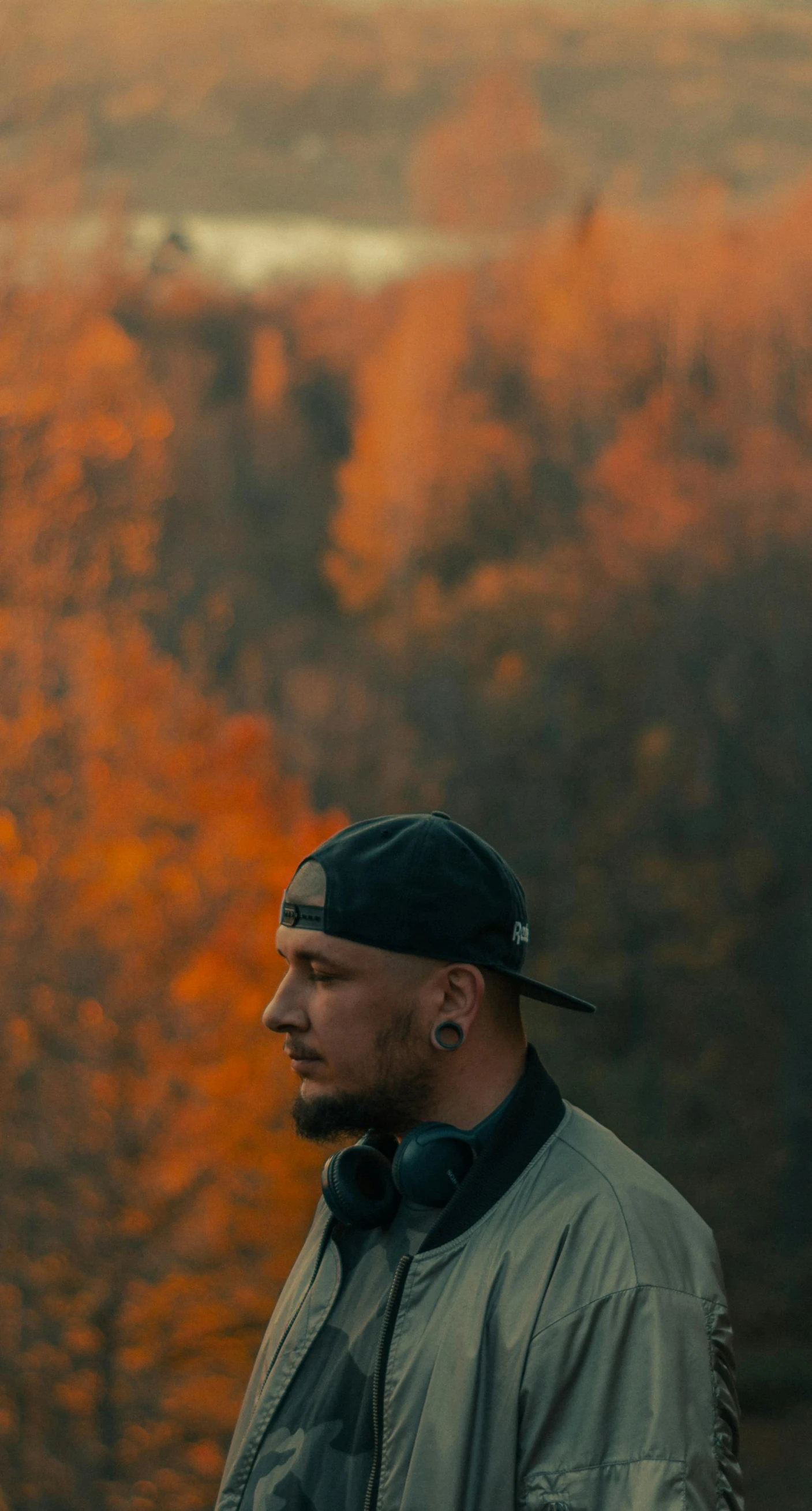 a man standing on top of a hill next to a forest, a photo, by Attila Meszlenyi, pexels contest winner, orange and brown leaves for hair, with rap cap on head, profile image, smokey